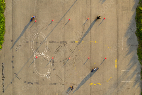 top down wide aerial view of four riders practicing on an advanced motorcycle training slalom course between orange cones with long shadows, painted lines and tire marks 