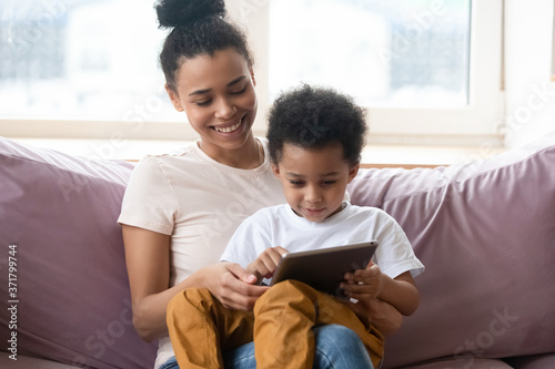 Once upon a time. African millennial mother sitting on sofa at living room holding toddler son on knees watching cartoon on tab pc screen, black nanny teaching little boy to read using education app