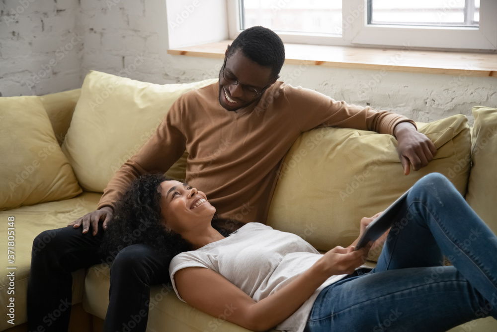 Just the two of us. Positive happy african couple in love spending weekend at home together, smiling black wife lying on husband knees on cozy couch at living room holding digital tablet pc computer