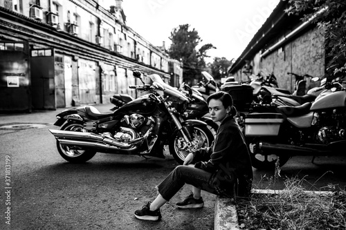A beautiful lovely girl sits on the curb near the old-fashioned motorcycles on the territory of the factory. Black and white.