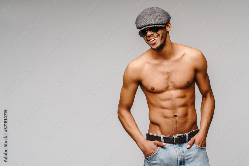 Sexy young muscular african american man shirtless wearing sunglasses and cap over light grey background