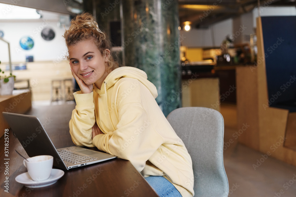 Smiling attractive woman sitting in cafe, drinking coffee and using laptop. Beautiful girl looking at camera happy while working remote from cafe. Female student studying outdoors using laptop