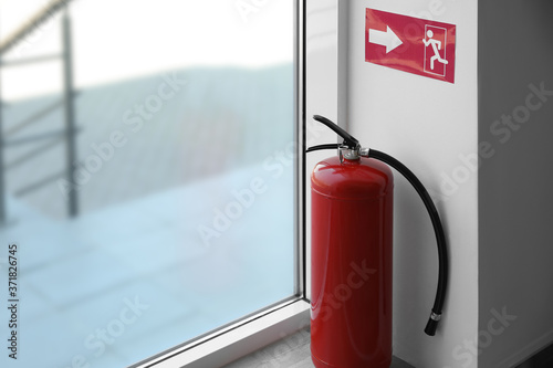 Modern fire extinguisher and emergency exit sign near window indoors Fototapeta