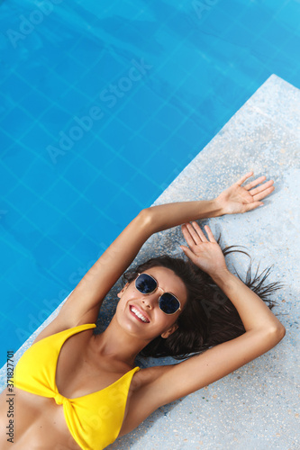 Top view of attractive smiling tanned woman enjoying sun. Sexy female sunbathing near pool edge, resting after swimming at poolside. Elegant lady tourist relax under sun of paradise hotel resort