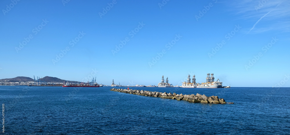 Large commercial harbour in the afternoon, ships, cranes, breakwaters and oilrigs, crisp horizon and hills visible in distant background.
