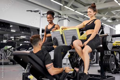 relax time in gym, two diverse women have talk with sportsman, they discuss something, smile, have rest, take a break during workout, weightlifting