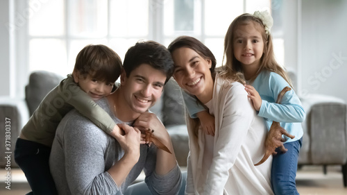Portrait of happy young mixed race woman with loving husband having fun with adorable little son and daughter, smiling nice family playing in living room, looking at camera, good relations concept.