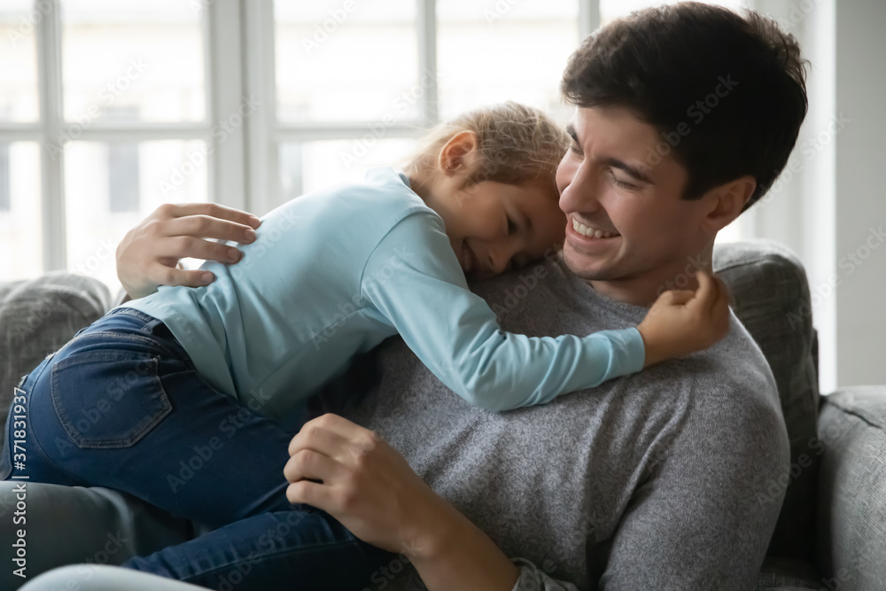 Affectionate little baby girl embracing smiling father, lying together on comfortable couch indoors. Caring happy young handsome daddy showing love and care to small cute child daughter at home. Affectionate little baby girl embracing smiling father, lying together on comfortable couch indoors. Caring happy young handsome daddy showing love and care to small cute child daughter at home.