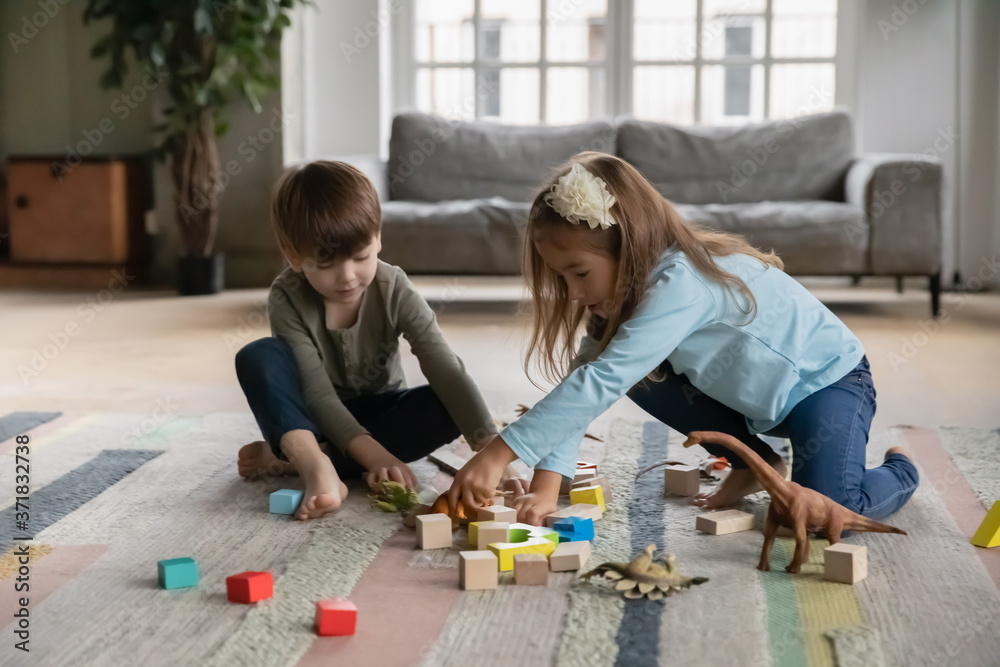 Naklejka premium Cheerful little kids siblings sitting on warm floor, enjoying playing with cubes and toys in living room. Happy small brother and sister best friends having fun together without parents indoors.