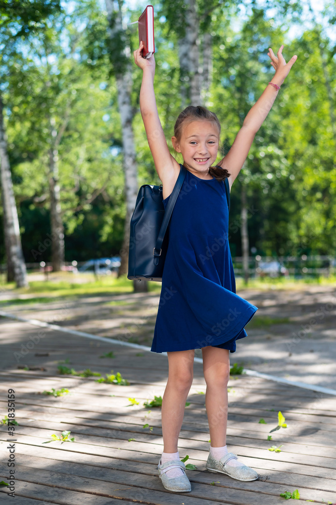 Excited schoolgirl is holding a textbook and jumping in the park. The girl is happy to go back to school.