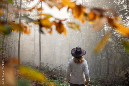Moody atmosphere in autumn forest. Woman wearing knitted sweater and hat standing in fog at woodland