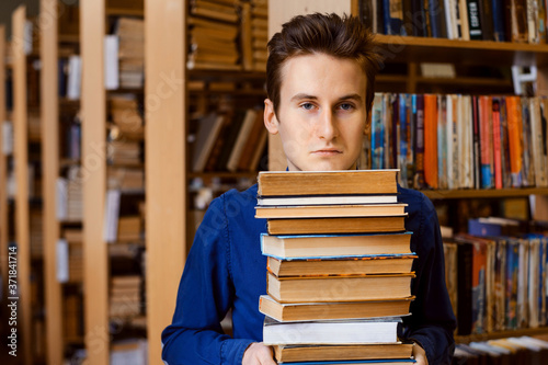 Male student stands with a pile of books at the library and looks to the camera with a very unhappy, tired and disappointed look.