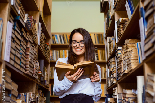 Beautiful brunette student girl stands at the library between shelves full of books and reads something with a smile on her face.