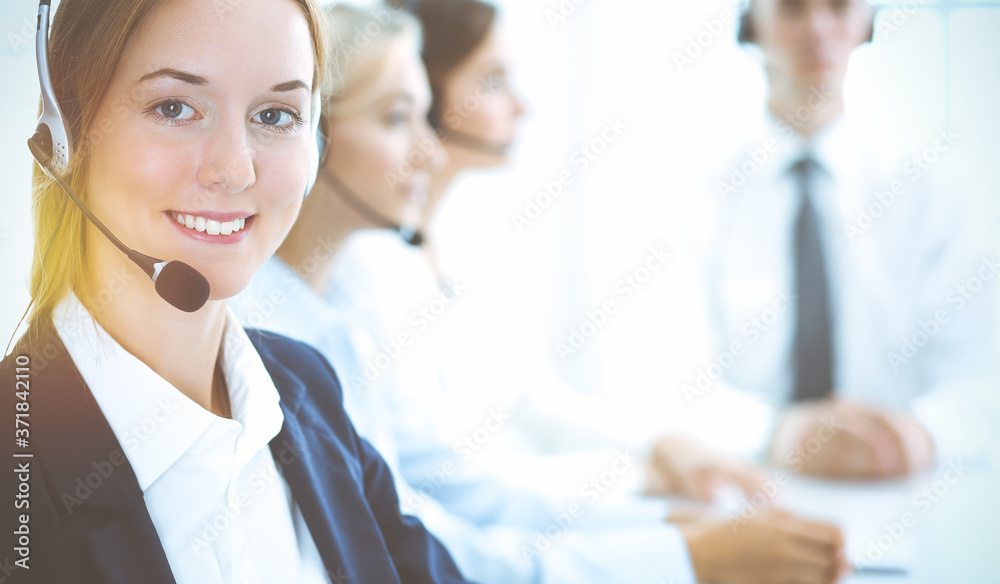 Cheerful smiling business woman with headphones consulting clients. Group of diverse phone operators at work in sunny office.Call center and business people concept Cheerful smiling business woman with headphones consulting clients. Group of diverse phone operators at work in sunny office.Call center and business people concept