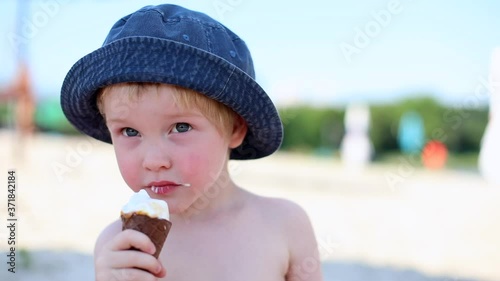 Front view of little cute happy blonde boy eating tasty ice-cream on sunny sandy beach with emotional face. Traveling kids. Fashion jeans panama hat. Male child enjoying vacations with family outdoor.