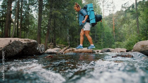 Woman tourist with backpack crossing stream in forest, travel adventure concept