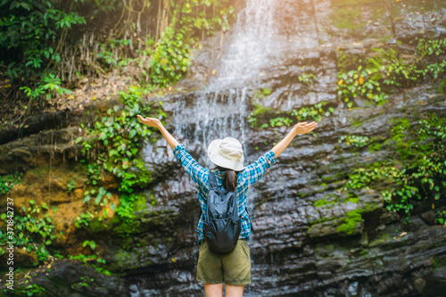 Fototapeta Naklejka Na Ścianę i Meble -  Asian girl tourist traveling adventure exploring nature jungle trail into forest waterfall open arms in success and peace, wearing travel gear bag hat, summer hot healthy outdoor freedom lifestyle