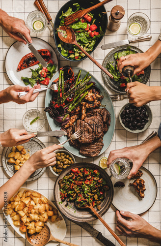 Summer barbeque party. Flat-lay of table with grilled meat, vegetables, salad, roasted potato and peoples hands over checkered white tablecloth, top view. Family gathering, comfort food concept