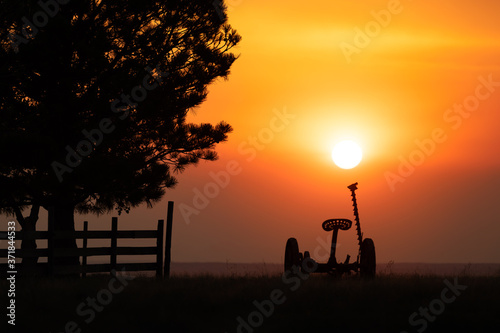 Forest fire smoke fills the air at sunset on a farm in eastern Colorado. The sky is very orange. This is a silhouette scene with a tractor and tree visible in the foreground.