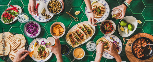 Family or friends home taco party. Flat-lay of Mexican traditional dishes Tacos with beef meat, corn tortillas , tomato salsa and peoples hands over green background, top view. Mexican cuisine concept photo