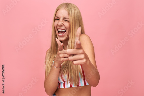 Cheerful excited hipster girl with loose straight hair winking, pointing fore fingers at camera, being in good mood. Stylish young Caucasian woman having fun, making gesture. Body language