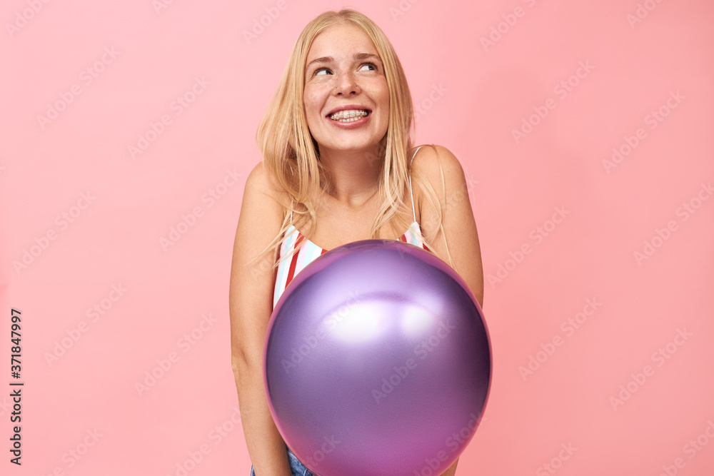 Studio image of beautiful shy teenage girl with long straight hair looking upwards with thoughtful smile, daydreaming posing isolated against blank pink copy space wall, holding helium balloon Studio image of beautiful shy teenage girl with long straight hair looking upwards with thoughtful smile, daydreaming posing isolated against blank pink copy space wall, holding helium balloon