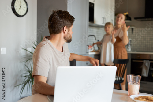 working man turned his head back, looks at his wife and daughter in the kitchen. freelance, work from home concept