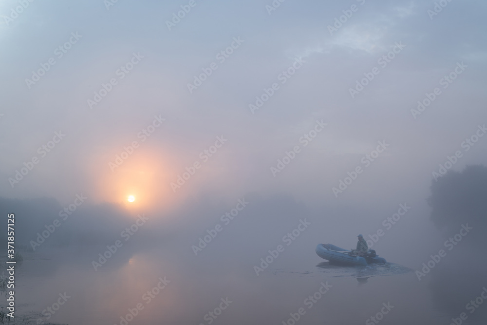 Tranquil morning landscape panorama at sunrise. A fisherman on a rubber boat on the river in heavy fog. Dawn illuminates and makes the clouds colorful.