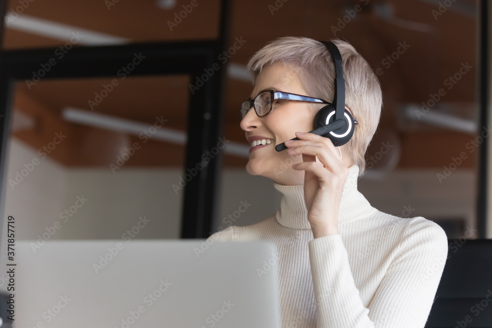 Happy young short haired 30s businesswoman in eyeglasses and headset with microphone consulting with colleague about decision, discussing contract or working issues with client online in office. Happy young short haired 30s businesswoman in eyeglasses and headset with microphone consulting with colleague about decision, discussing contract or working issues with client online in office.