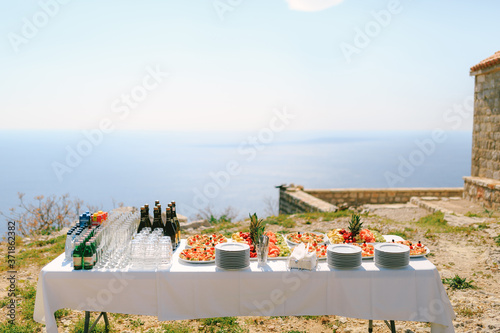 A festively served buffet table with water, champagne and fruits on plates against the backdrop of an old building by the water.