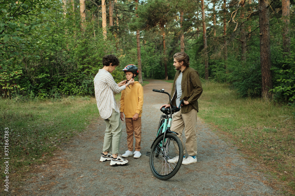 Young careful mother helping her son with tying up belts of sports helmet Young careful mother helping her son with tying up belts of sports helmet