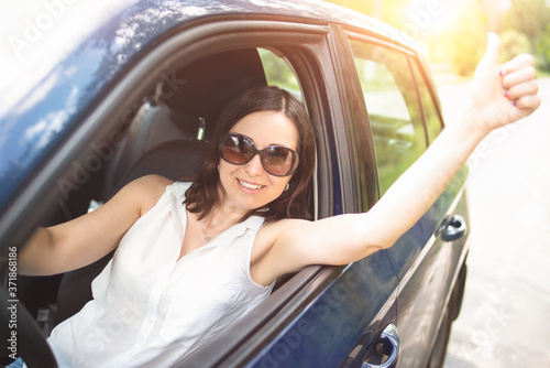 A smart looking middle-aged adult woman wearing sunglasses put her hand out the car window and raised her thumbs up. smiling woman drives Car