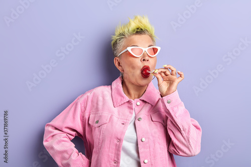 beautiful glamour senior woman in sunglasses licking chupa chups isolated over blue background. lifestyle, free time photo