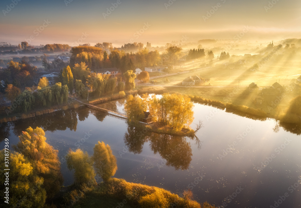Aerial view of house with colorful trees on small island on the lake. Foggy sunrise in autumn. Beautiful landscape with village in fog, golden sunbeams, reflection in water. Fall in Ukraine. Top view