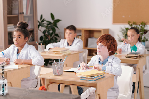 Group of intercultural elementary schoolkids in whitecoats sitting by desks