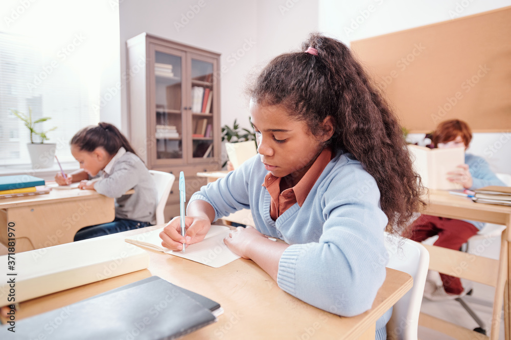 Serious mixed-race schoolgirl making notes or wtiting essay in copybook Serious mixed-race schoolgirl making notes or wtiting essay in copybook