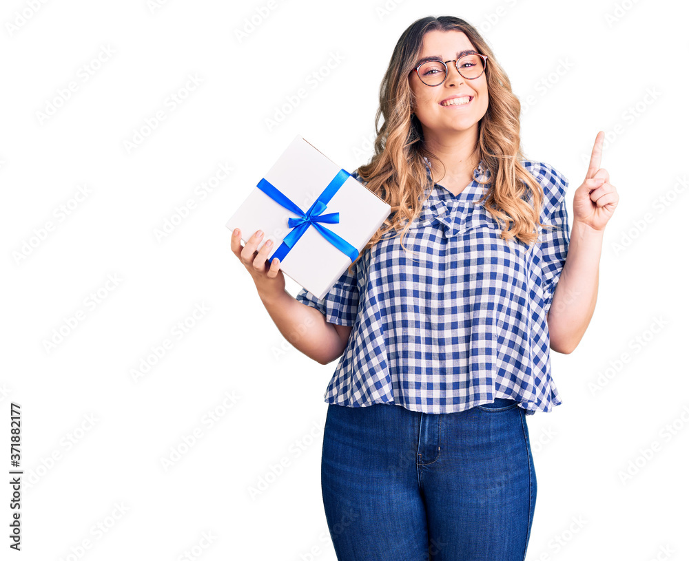 Young caucasian woman holding gift surprised with an idea or question pointing finger with happy face, number one Young caucasian woman holding gift surprised with an idea or question pointing finger with happy face, number one