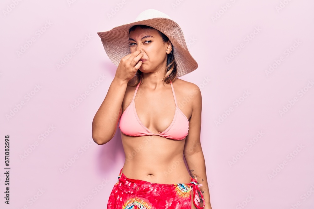 Young woman wearing bikini and hat smelling something stinky and disgusting, intolerable smell, holding breath with fingers on nose. bad smell Young woman wearing bikini and hat smelling something stinky and disgusting, intolerable smell, holding breath with fingers on nose. bad smell