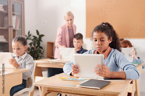 Group of youthful contemporary schoolchildren with touchpads sitting by desks