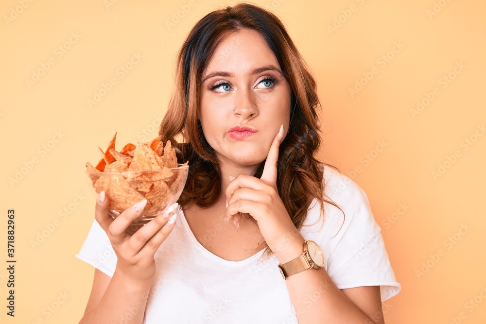 Young beautiful caucasian woman holding nachos potato chips serious face thinking about question with hand on chin, thoughtful about confusing idea Young beautiful caucasian woman holding nachos potato chips serious face thinking about question with hand on chin, thoughtful about confusing idea