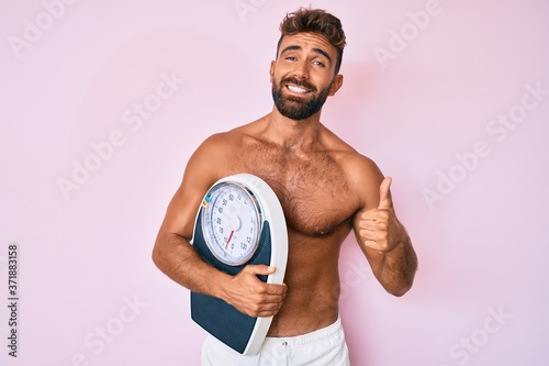 Young hispanic man standing shirtless holding weighing machine smiling happy and positive  thumb up doing excellent and approval sign