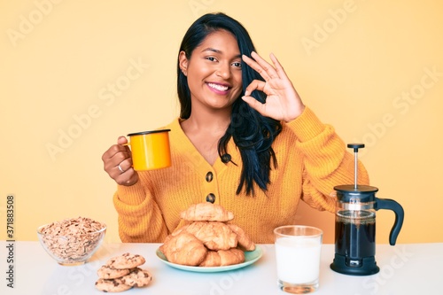 Beautiful latin young woman with long hair sitting on the table having breakfast doing ok sign with fingers  smiling friendly gesturing excellent symbol