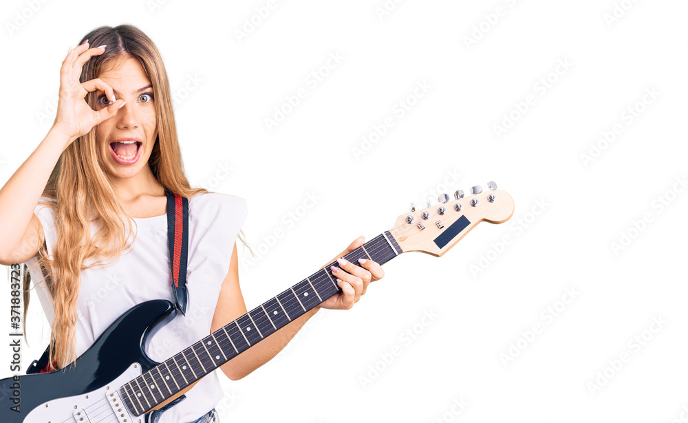 Fototapeta premium Beautiful caucasian woman with blonde hair playing electric guitar smiling happy doing ok sign with hand on eye looking through fingers