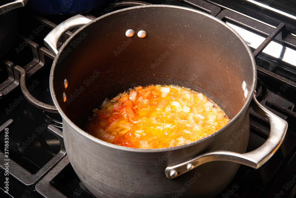 Black pot on stove with oil, onions and garlic adding tomatoes preparing a meal Black pot on stove with oil, onions and garlic adding tomatoes preparing a meal
