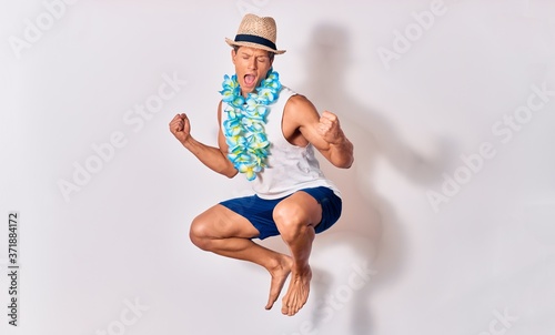 Young handsome hispanic tourist man on vacation wearing summer clothes and hawaiian lei smiling happy. Jumping with smile on face celebrating with fists up over isolated white background