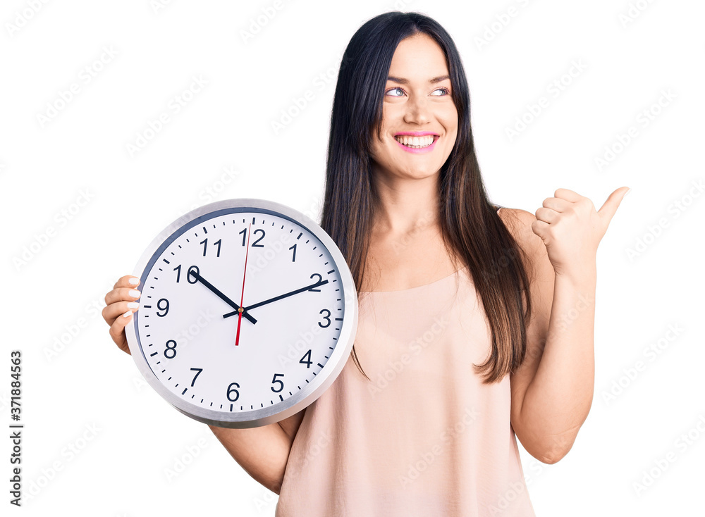 Young beautiful caucasian woman holding big clock pointing thumb up to the side smiling happy with open mouth Young beautiful caucasian woman holding big clock pointing thumb up to the side smiling happy with open mouth