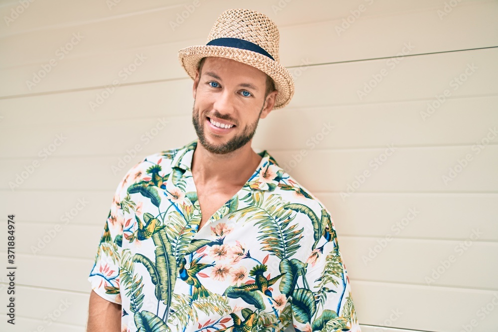 Handsome caucasian man wearing summer hat and flowers shirt smiling happy leaning on the wall Handsome caucasian man wearing summer hat and flowers shirt smiling happy leaning on the wall