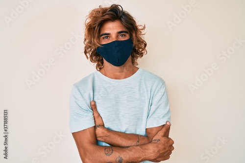 Young hispanic man wearing covid-19 mask happy face smiling with crossed arms looking at the camera. positive person.