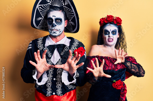 Young couple wearing mexican day of the dead costume over yellow disgusted expression, displeased and fearful doing disgust face because aversion reaction.