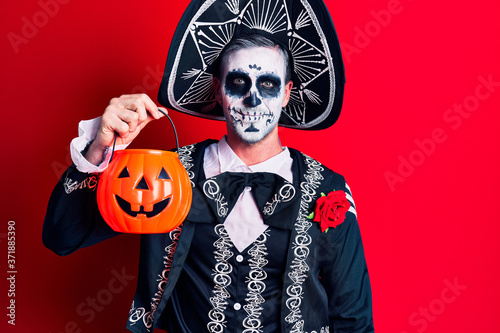 Young man wearing mexican day of the dead costume holding pumpkin looking positive and happy standing and smiling with a confident smile showing teeth